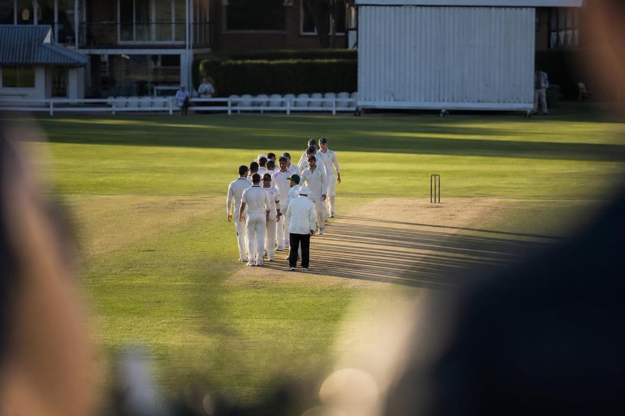 two teams in whites shaking hands at the end of a match, seen from a distance in the middle of a green ground, long afternoon shadows, no faces visible. (AI-generated image)