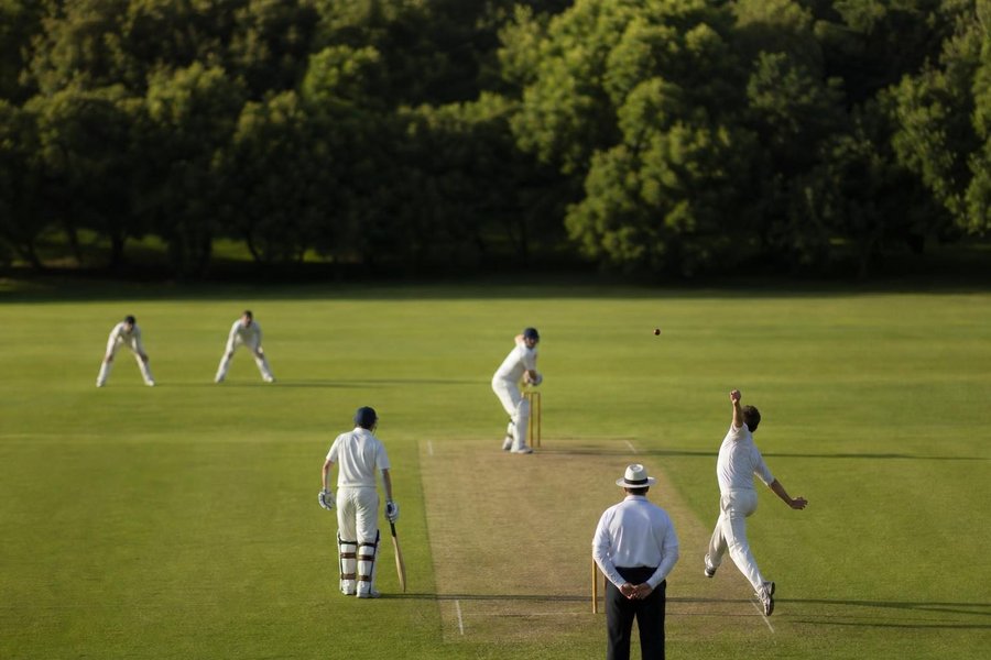 A bowler in delivery stride seen from the side at a long distance, the batter ready at the crease, an even green ground, strong contrast. (AI-generated image)