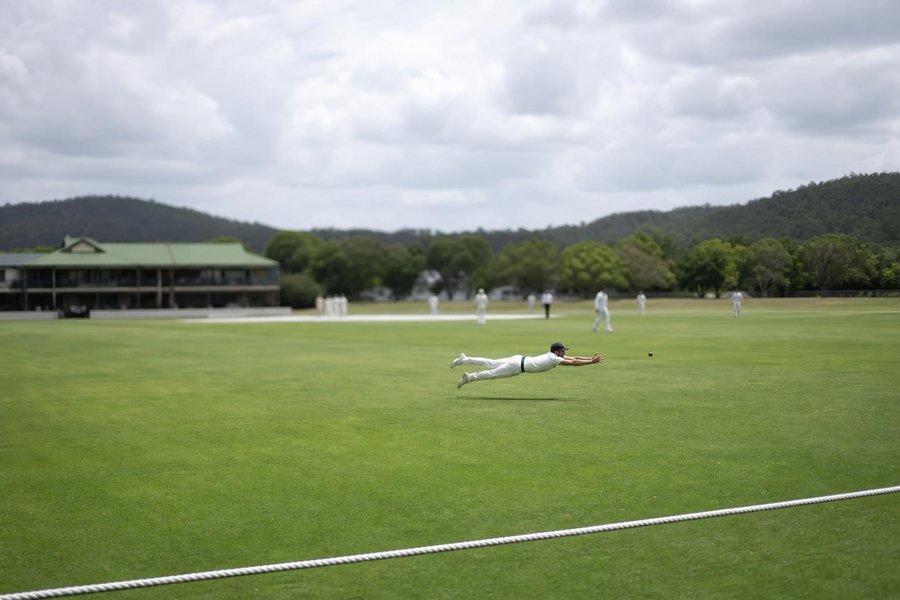 A cricket fielder diving to take a catch at square leg on a cricket oval with a white rope boundary, the cricket pitch and a pavilion visible behind, frozen motion, small in the frame. (AI-generated image)