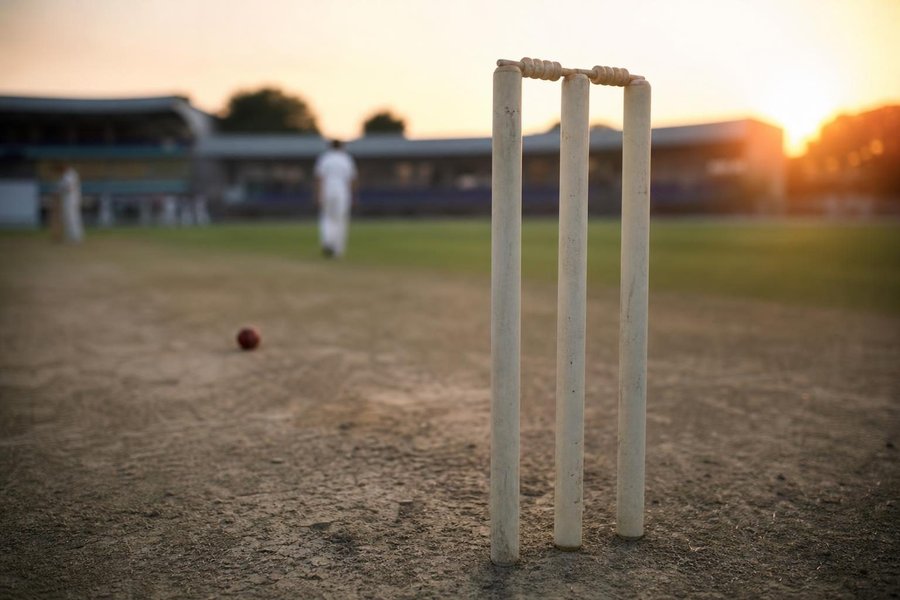 stumps close-up at dusk with a ball just beyond, warm backlight, a faint blur of a stadium behind. (AI-generated image)