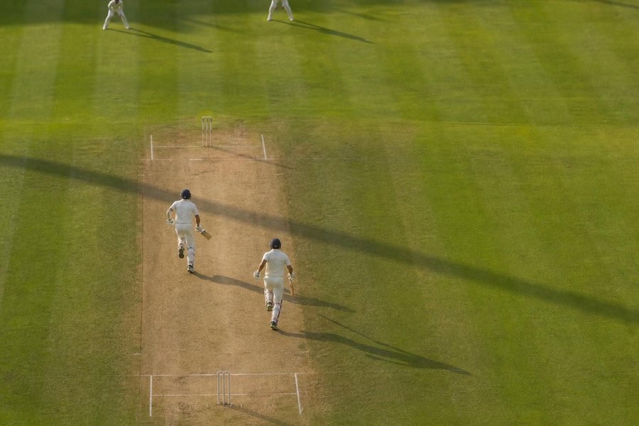 two batters running between the wickets, seen from high above in a wide shot, long shadows across the pitch. (AI-generated image)