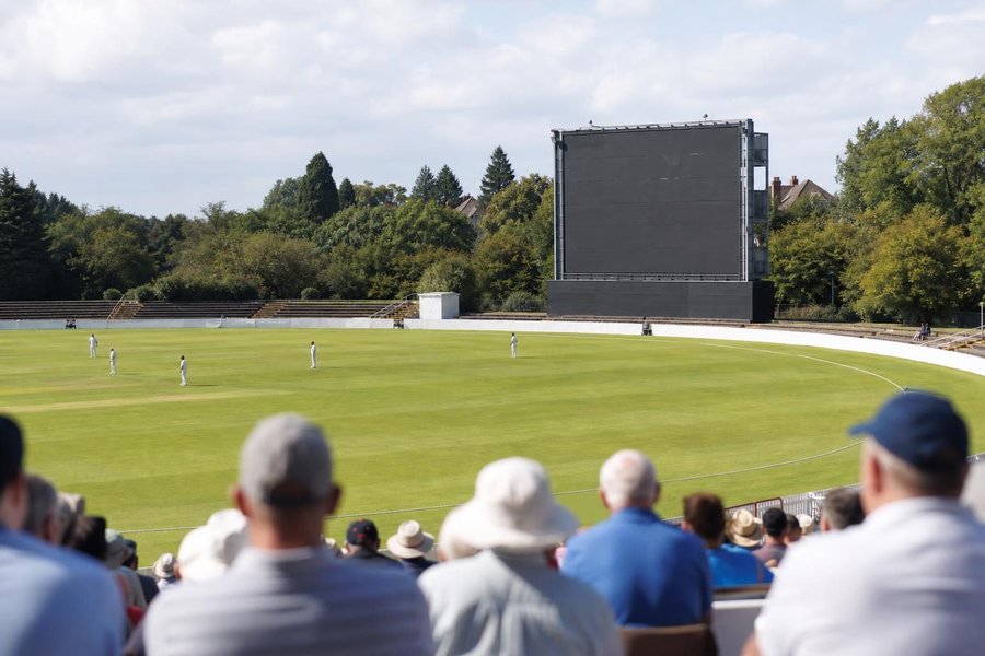 A traditional cricket ground seen from the stands, a large blank manual scoreboard tower at the boundary, spectators in the foreground from behind, sunlit. (AI-generated image)