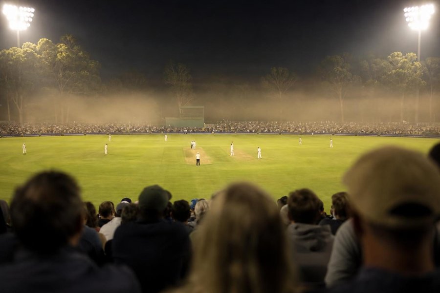An Australian cricket stadium on a summer night under floodlights, the crowd in the stands, a bowler in the run-up seen far away, warm haze over the outfield, gum trees outside the ground. (AI-generated image)