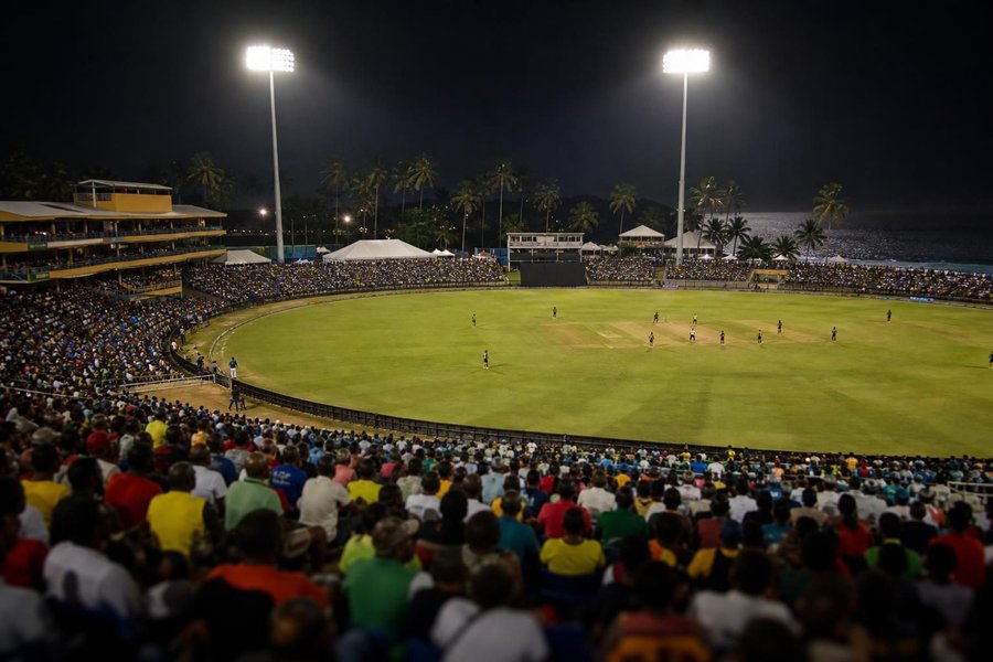 A Caribbean cricket ground at night under lights, palm trees beyond the stands, a lively crowd in bright clothes, the sea faintly visible at the edge of the ground. (AI-generated image)