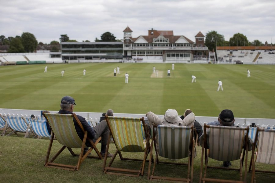 A traditional English county cricket ground with a large pavilion, a four-day match in progress, a handful of spectators in deckchairs, grey-white sky. (AI-generated image)