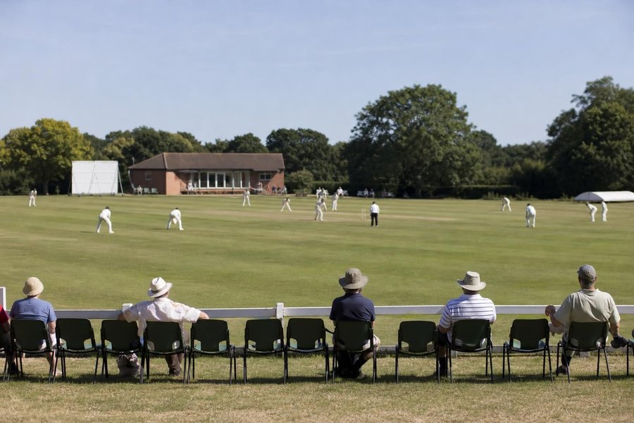 A smaller county ground with a modest brick pavilion and trees, a four-day match, a few spectators, wide shot in soft sunshine. (AI-generated image)