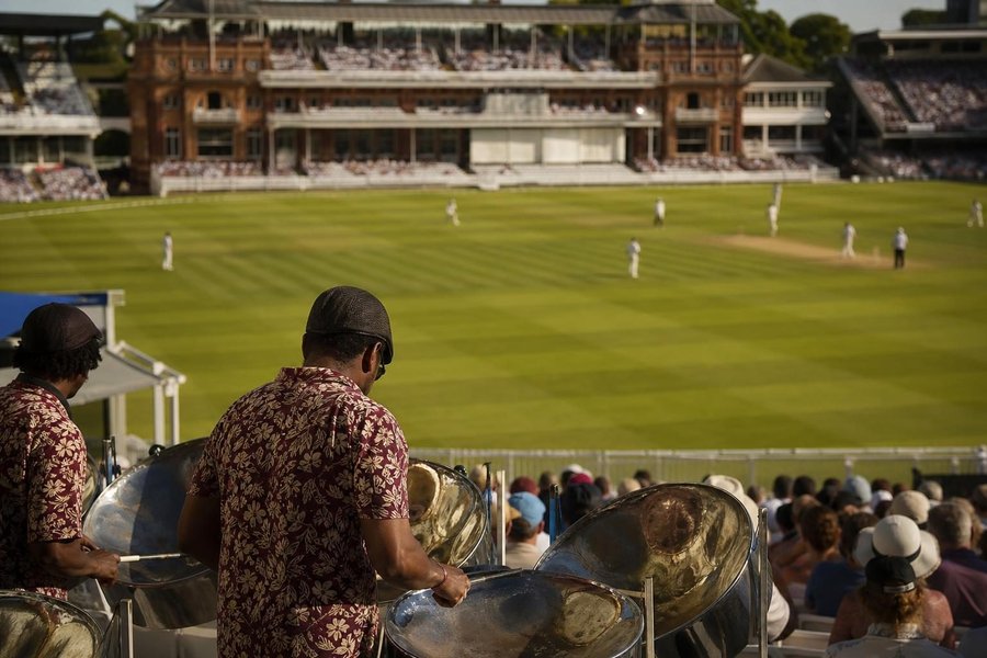A summer Test match at an English ground, a West Indian-style steel band and fans in the stands, a bowler running in far away, warm light. (AI-generated image)