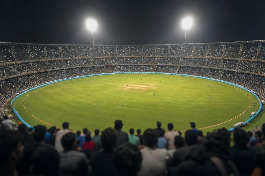 An enormous stadium packed with spectators in the evening, thousands of lights and the glow of floodlights, a batter small on the pitch, seen from the upper tier. (AI-generated image)