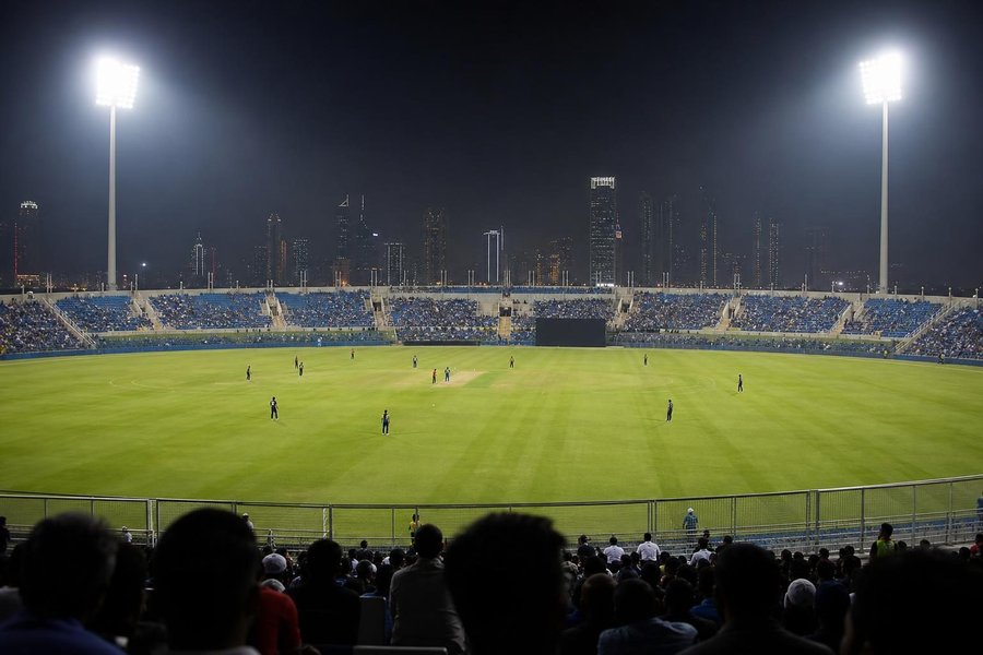 A modern desert-country cricket stadium at night under floodlights, even green outfield, rows of seats with a modest crowd, a hazy skyline far behind. (AI-generated image)