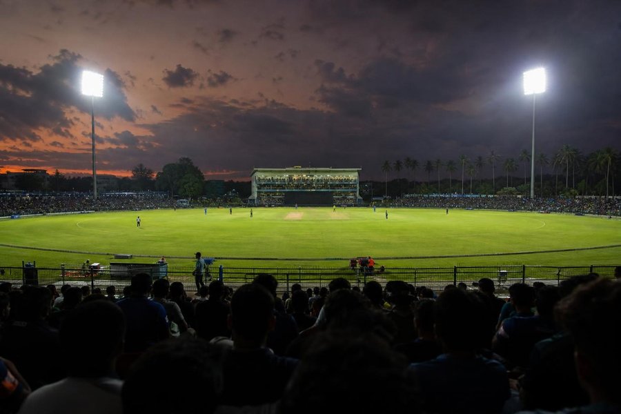 A cricket stadium in Sri Lanka at night under floodlights, tropical dusk sky, palm trees outside the stands, a wide shot of a night match in progress. (AI-generated image)