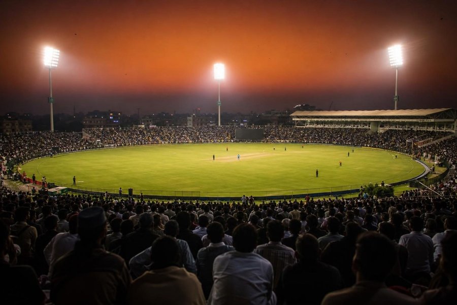 A floodlit stadium in Pakistan at dusk during a night match, a dense crowd in the stands, a hazy orange sky above the floodlight towers, a batter small in the middle. (AI-generated image)