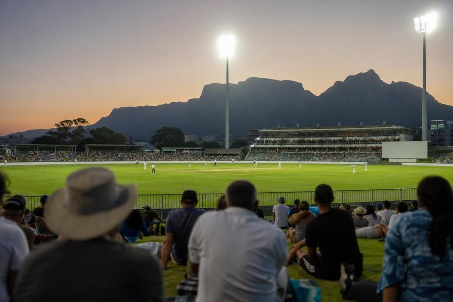 A South African cricket stadium at dusk, floodlights coming on, a mountain silhouette behind the stands, a crowd on grass banks, a bowler in his delivery stride far away. (AI-generated image)