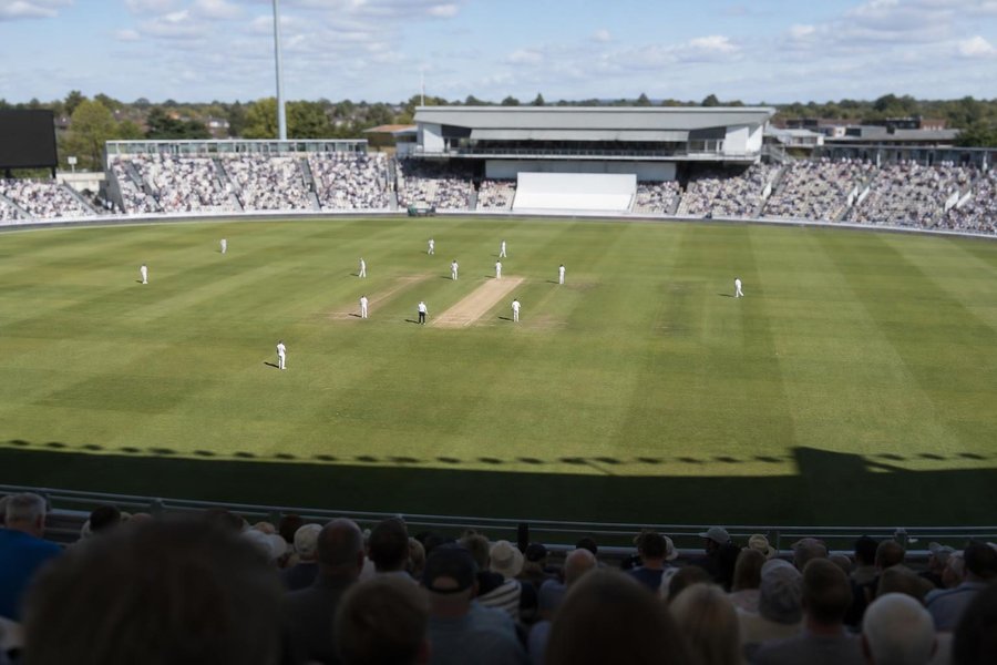 A five-day Test match at a big ground, players in white in the middle, red ball and the long shadow of a pavilion, a quiet crowd, bright daylight. (AI-generated image)
