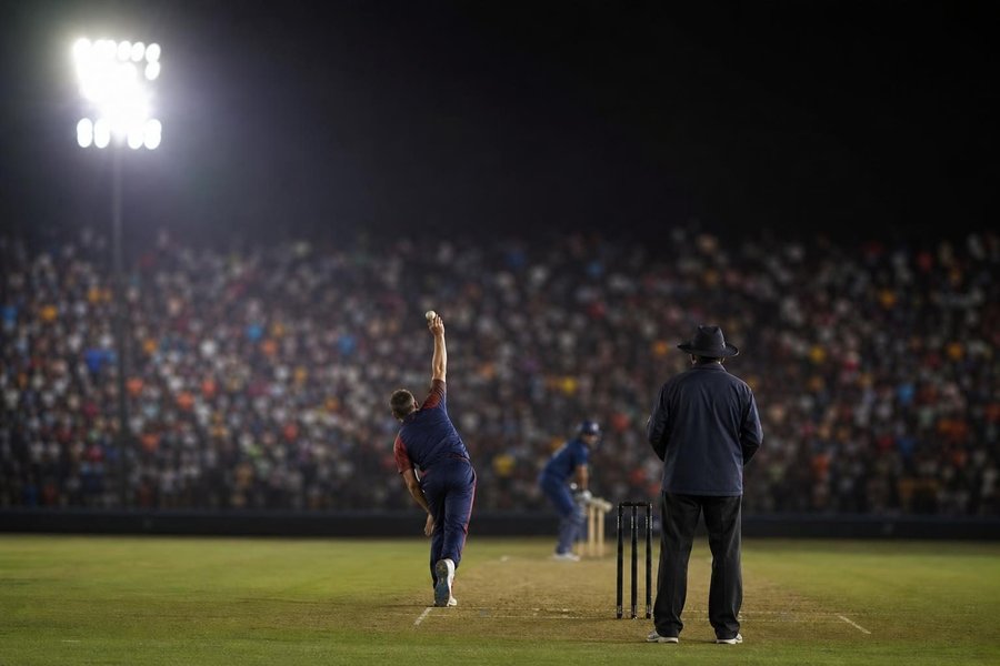 A London cricket ground at night under lights with a youthful, noisy crowd, a bowler about to deliver, bright but blurred crowd colours. (AI-generated image)