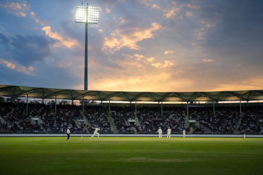 An English county ground on a summer evening, lively crowd in the stands, floodlights just switching on, a bowler in his run-up seen from side-on and far away. (AI-generated image)