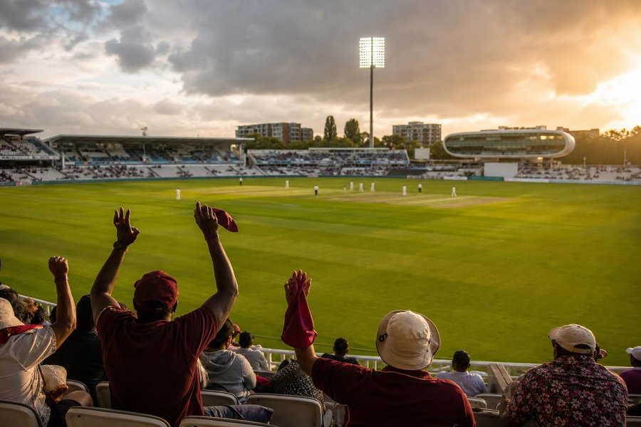 caribbean fans waving in the stands of an English ground, green outfield, a pavilion and rain clouds breaking, golden light. (AI-generated image)