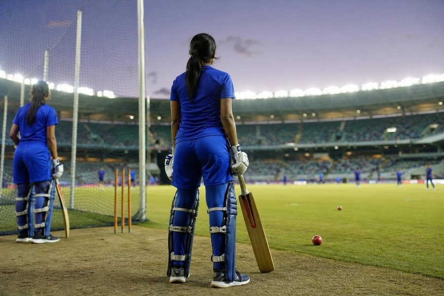 A cricket team of women in matching blue kit warming up with cricket bats and a red cricket ball at the edge of a floodlit cricket oval, stumps in a net behind them, a stadium of spectators beyond, no faces visible. (AI-generated image)