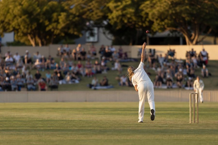 A women's cricket match at an Australian ground on a summer evening, a bowler in follow-through seen from behind and far away, spectators on grass banks, warm light. (AI-generated image)