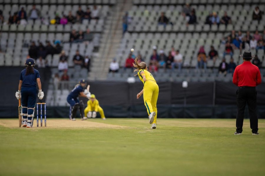 A women's international one-day match underway, bowler in follow-through seen from behind and far away, green outfield, stands partly full, daylight. (AI-generated image)