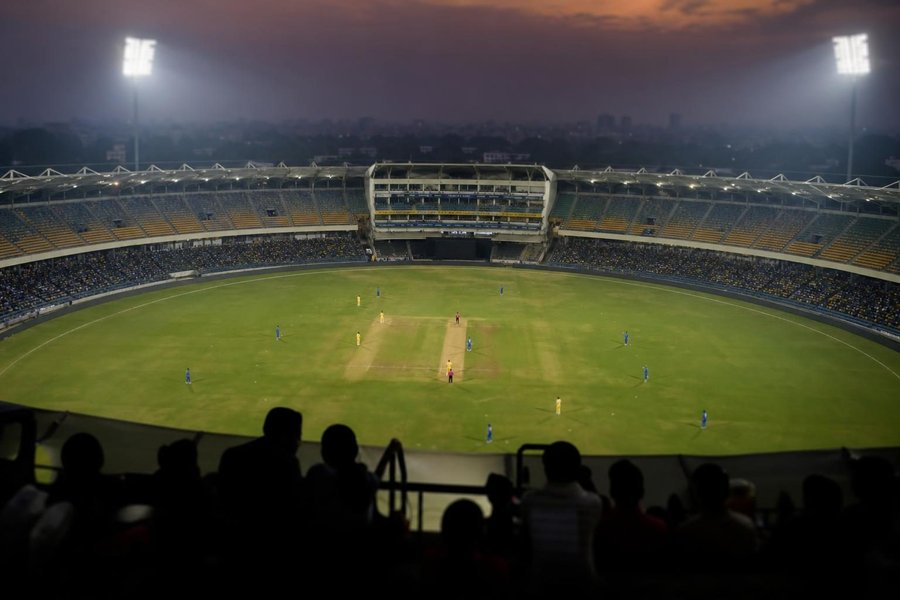 A women's cricket match at a large floodlit stadium in India at dusk, players in team colours seen from high in the stands, soft evening haze, no faces visible. (AI-generated image)