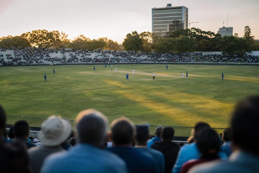 two cricket teams in white and in blue playing a one-day match on a cricket oval in Harare, seen from the stands far away, the pitch with stumps clearly visible at the centre, evening sun, faces not visible. (AI-generated image)