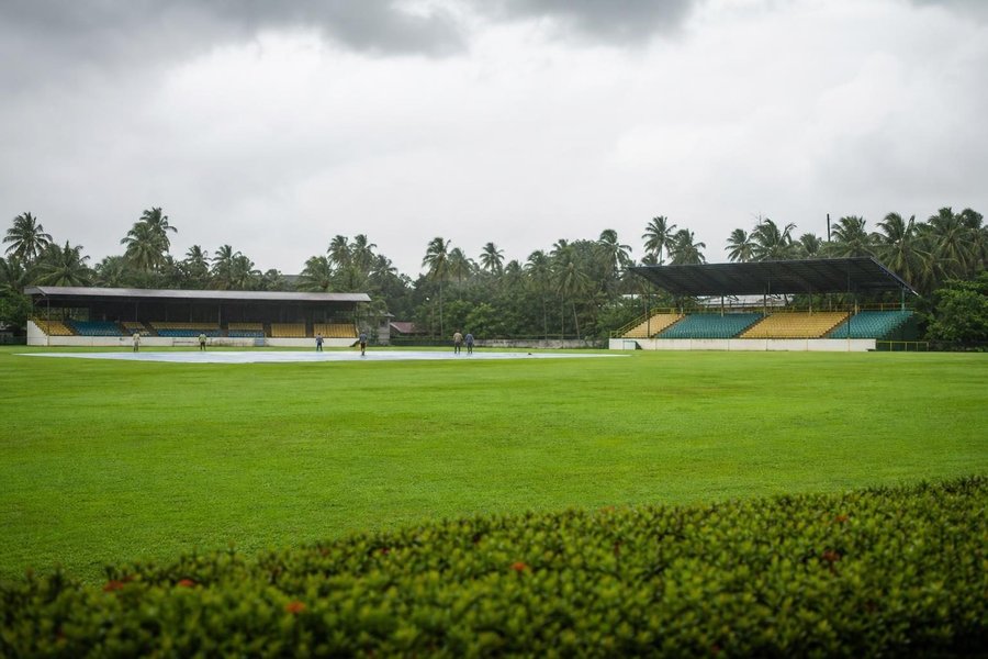 A cricket ground in humid monsoon light, lush green outfield after rain, heavy clouds, simple covered stands and palms at the edge. (AI-generated image)
