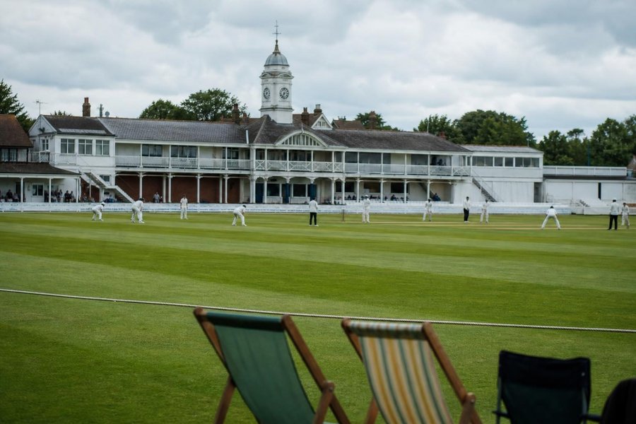 An English county cricket ground under soft overcast light, white pavilion with a clock tower, deckchairs and a rope boundary, freshly mown stripes. (AI-generated image)