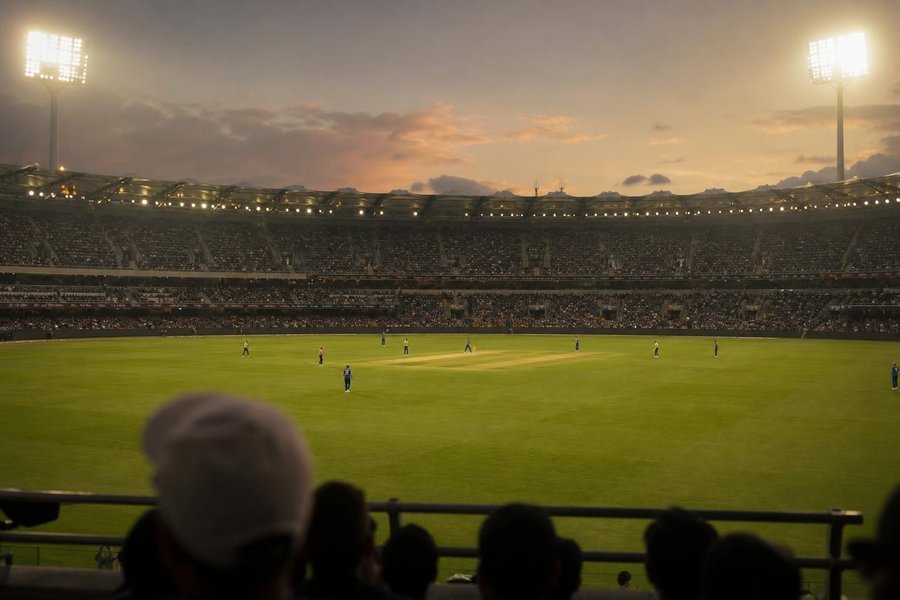 A huge floodlit cricket stadium at dusk, tiered stands full of spectators in warm haze, the floodlights glowing, a batter small in the centre. (AI-generated image)