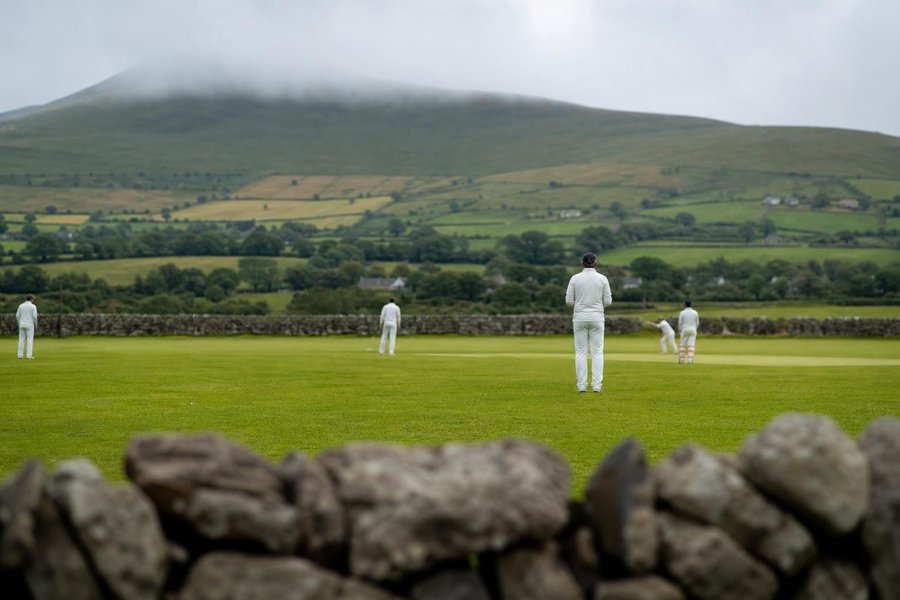 A small green cricket ground in Ireland, a low stone wall and misty rolling hills behind, soft rain-washed light, a few fielders in the distance. (AI-generated image)