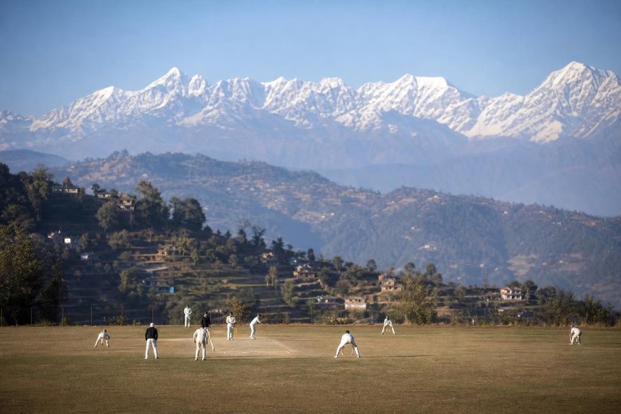 A cricket ground in the Kathmandu valley with snow-capped Himalayan peaks on the horizon, terraced hills, clear morning light, players in the distance. (AI-generated image)