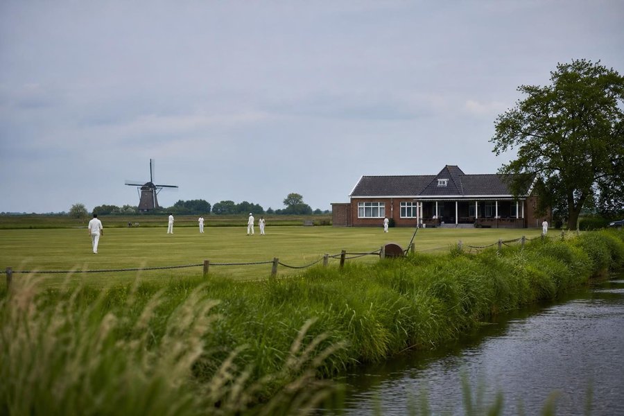 A club cricket ground in the Netherlands beside a canal, flat green land, a brick pavilion and a distant windmill, soft grey-blue sky. (AI-generated image)