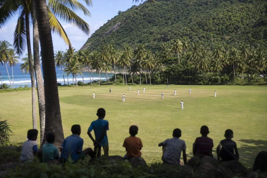 A tropical cricket ground beside the sea, coconut palms along the boundary, humid bright light, jungle-covered hills behind, children watching from the edge. (AI-generated image)