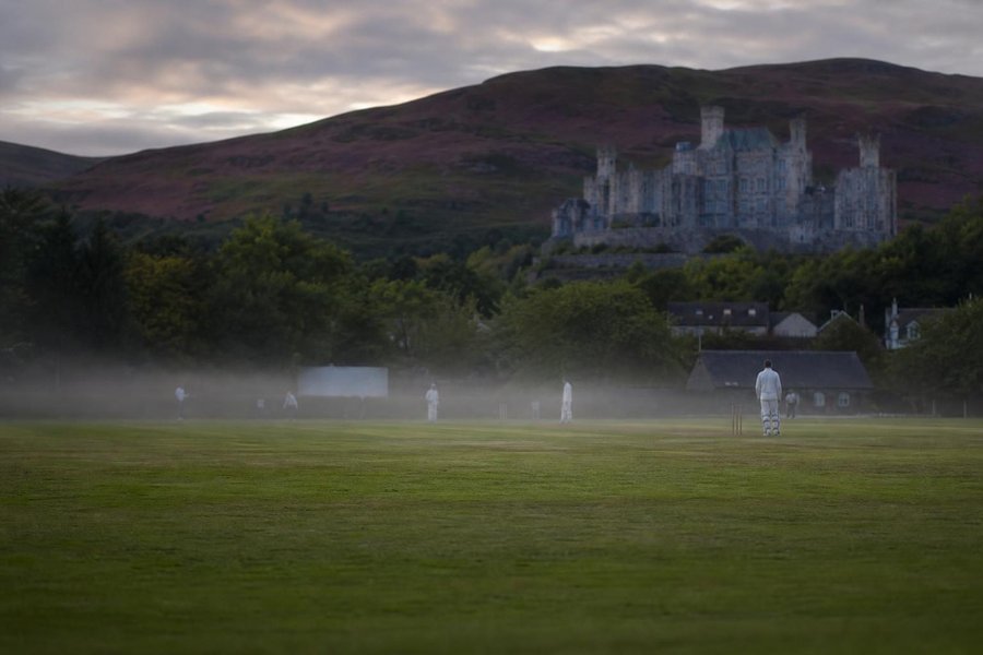 A Scottish cricket ground with a grey stone castle and heather hills behind it, cool low light, a thin mist over the outfield. (AI-generated image)
