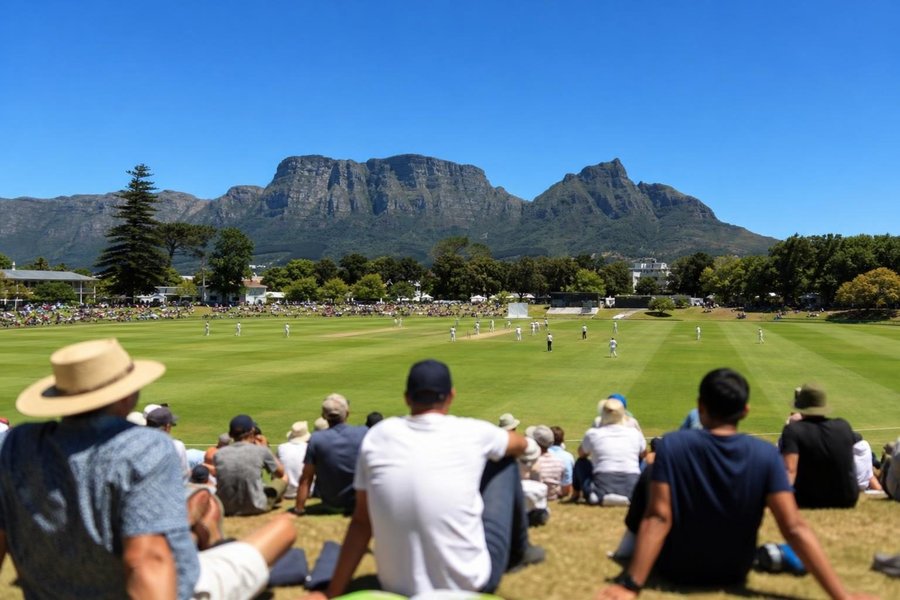 A cricket ground under a flat-topped mountain, intense blue sky, green outfield, a crowd on grassy banks, strong afternoon sun. (AI-generated image)
