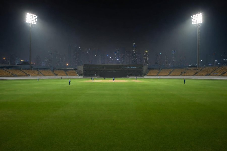 A modern cricket stadium in the desert at night under floodlights, a hazy city skyline far behind, perfectly even green outfield. (AI-generated image)