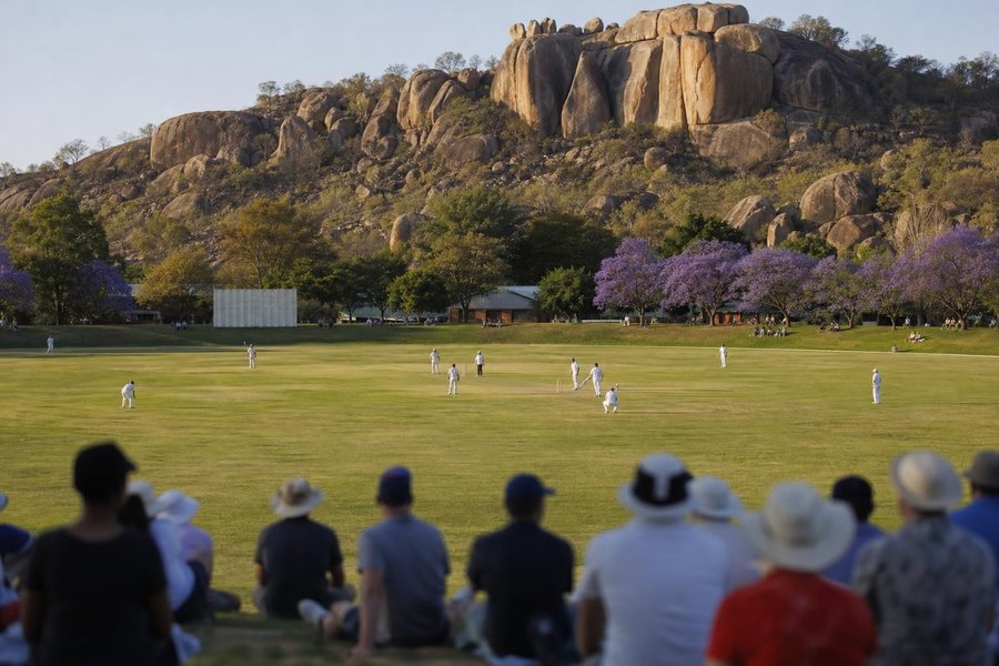 A cricket ground with large granite boulders rising behind the boundary, savannah light, jacaranda trees, a small crowd on the grass banks. (AI-generated image)